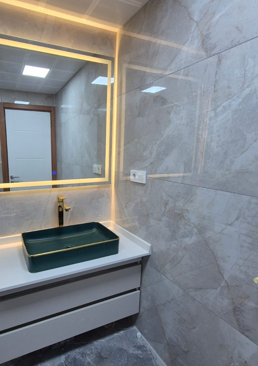 Altın çerçeveli ışıklı ayna, koyu yeşil tezgah üstü lavabo ve mermer duvarlı banyo